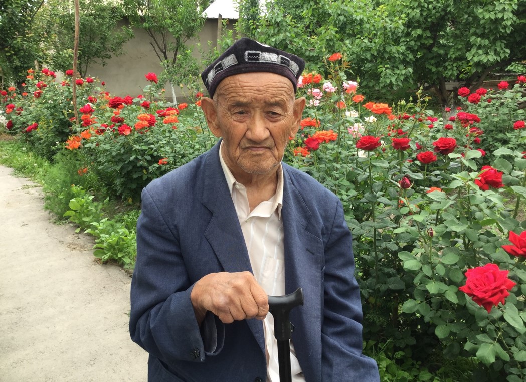 A photograph of an old Uyghur man standing in front of roses.